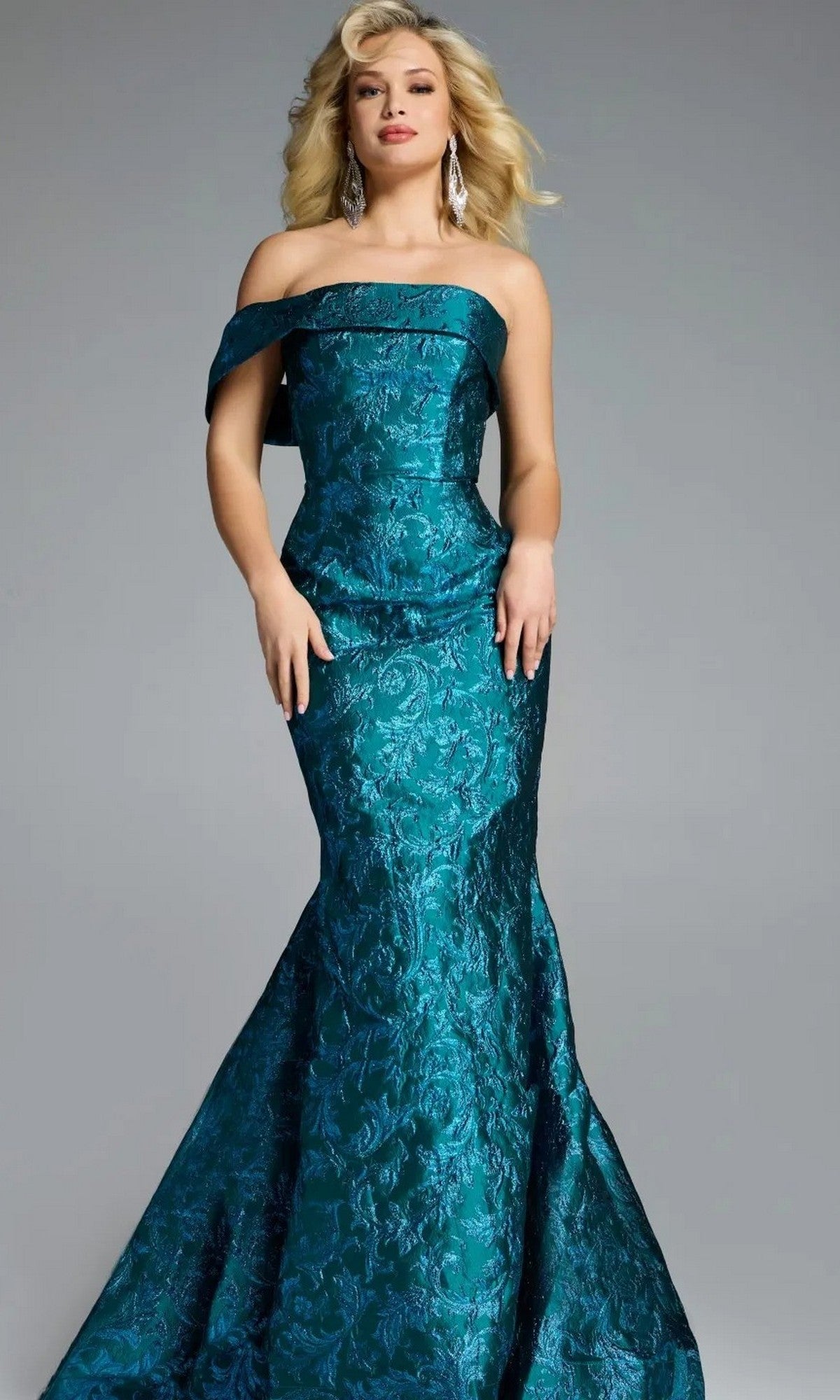 A woman wearing a Jovani 43978 long prom dress in peacock, featuring an off-shoulder neckline and floor-length textured floral design, stands against a plain gray background.